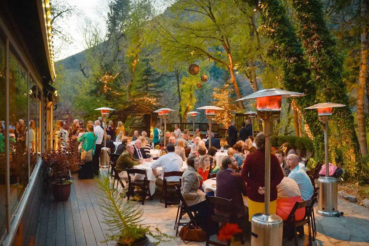 millcreek-img A group of people seated at outdoor tables, dining and socializing at an evening event surrounded by greenery and string lights.