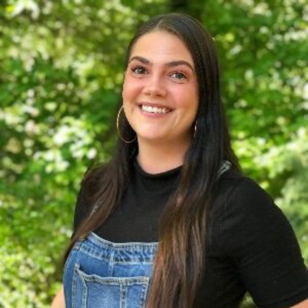 Untitled design (1) copy A woman with long dark hair wearing a black top and denim overalls smiles outdoors with a green leafy background.