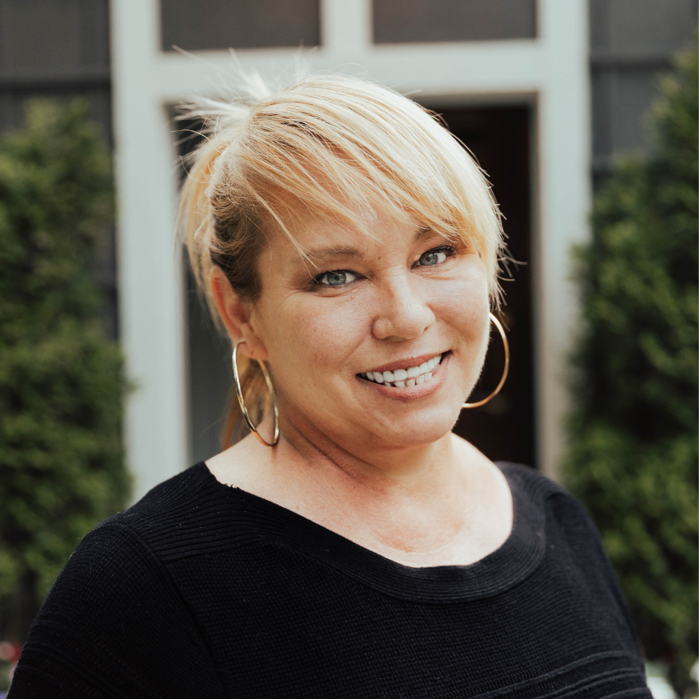 Untitled design (1) A woman with blonde hair, wearing large hoop earrings and a black top, smiles in front of a door and some greenery.